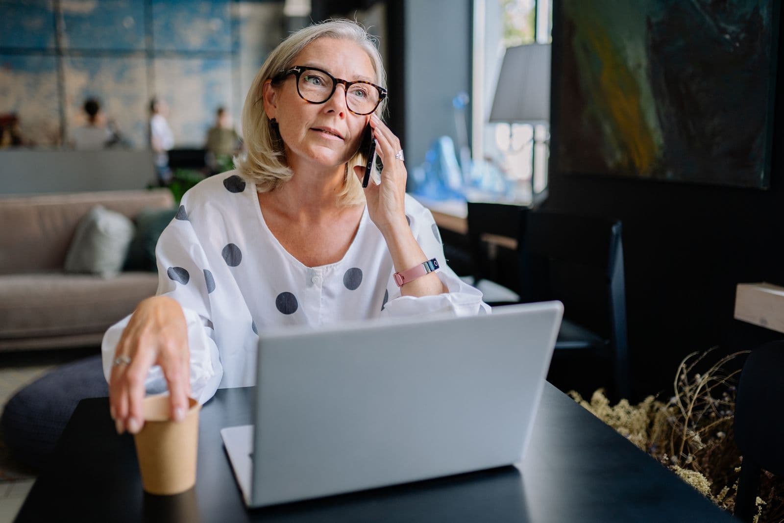Woman with glasses taking a phone call beside her laptop at a café table