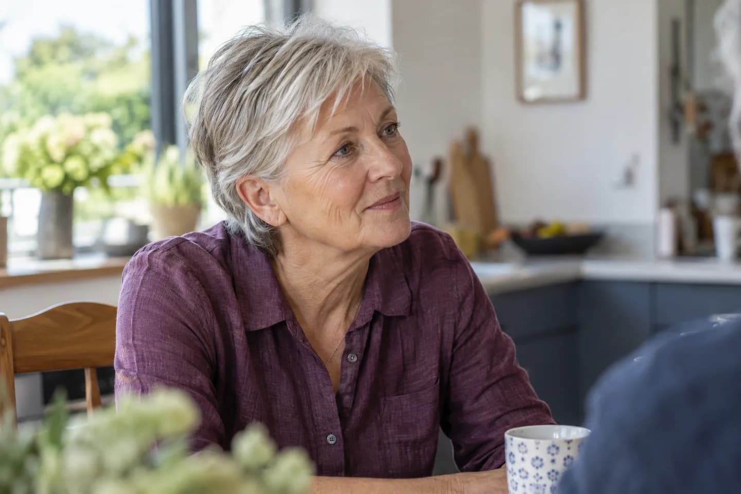 A silver-haired woman listening to a conversation at a bright kitchen table