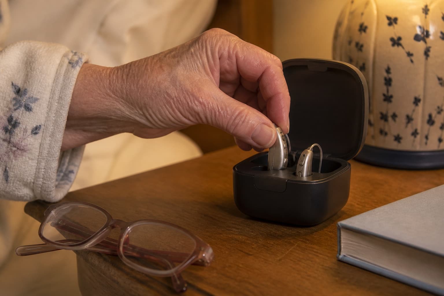Older woman placing two hearing aids into their charger on a bedside table