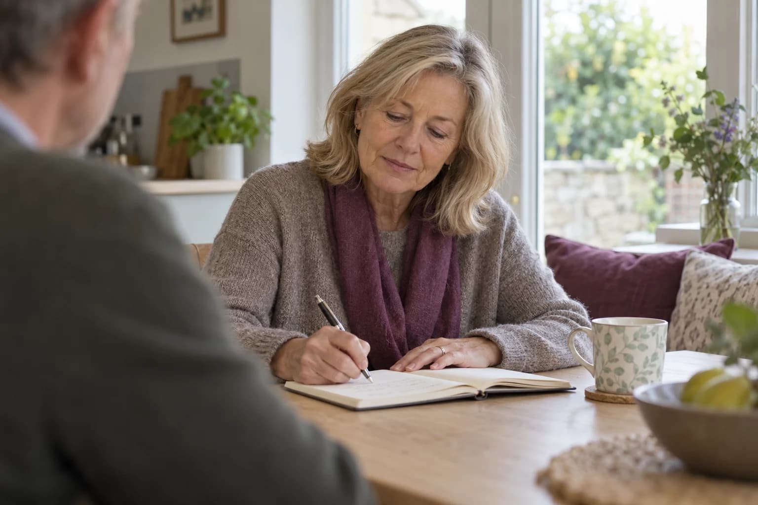 A woman writing notes in a bright home while a companion sits nearby